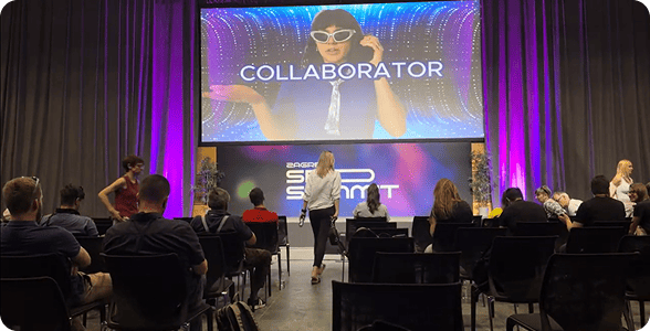 Участники конференции в Загребе, сидящие на стульях и смотрящие на большой экран, на котором показывается промо-видео Collaborator.